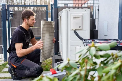 Instalator klęczy przy jednostce zewnętrznej pompy ciepła i wyjmuje filtr powietrza do kontroli